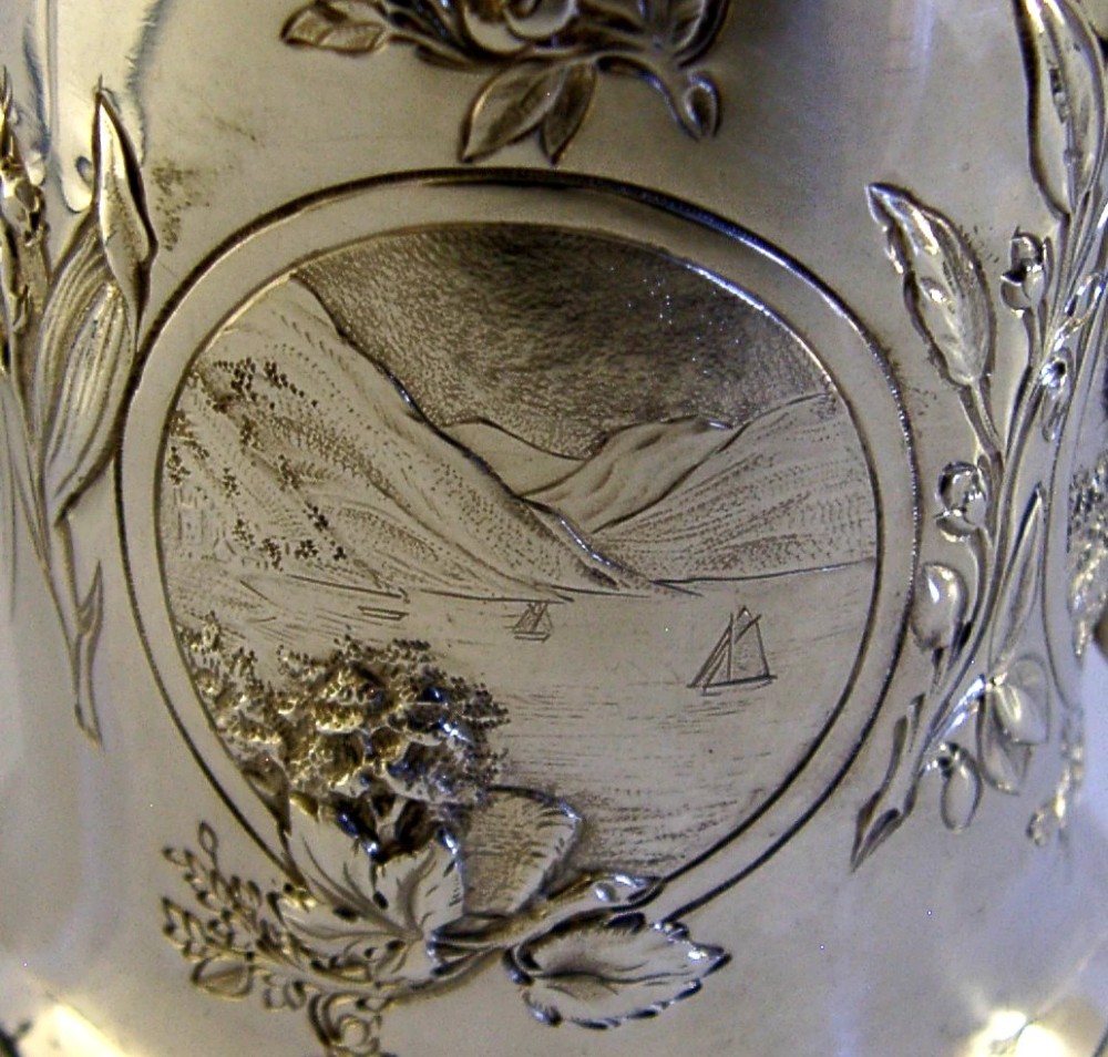 A Victorian sterling silver tankard mug featuring detailed relief floral decoration, a landscape roundel, a scroll handle, and a gilt interior. View V