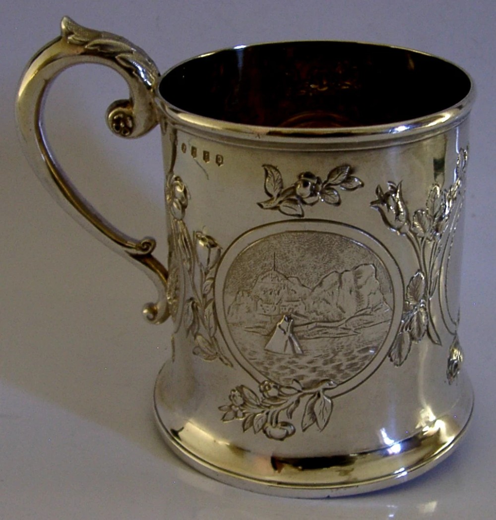 A Victorian sterling silver tankard mug featuring detailed relief floral decoration, a landscape roundel, a scroll handle, and a gilt interior. View VII