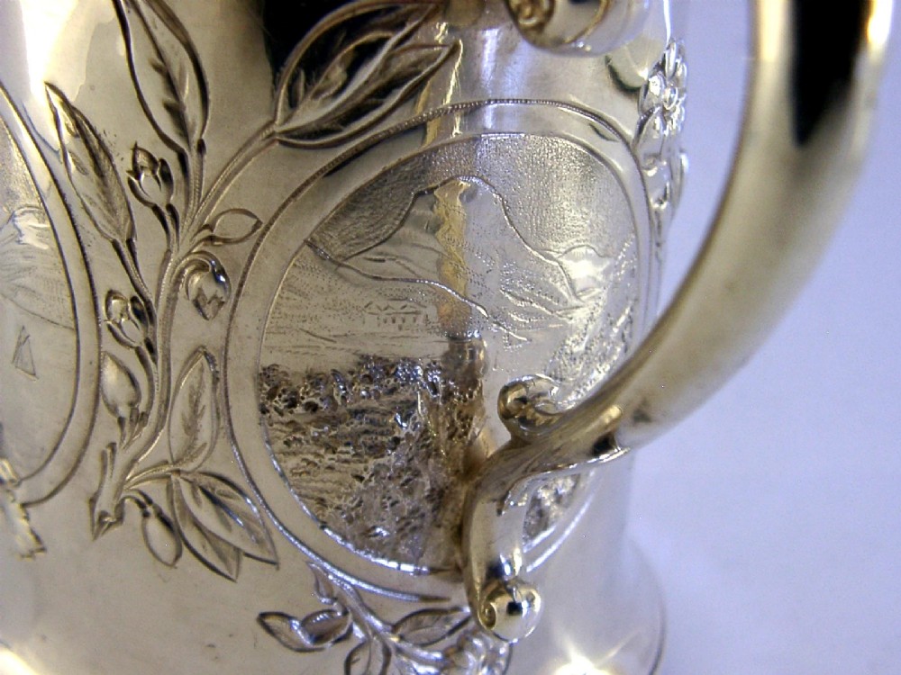 A Victorian sterling silver tankard mug featuring detailed relief floral decoration, a landscape roundel, a scroll handle, and a gilt interior. View IX
