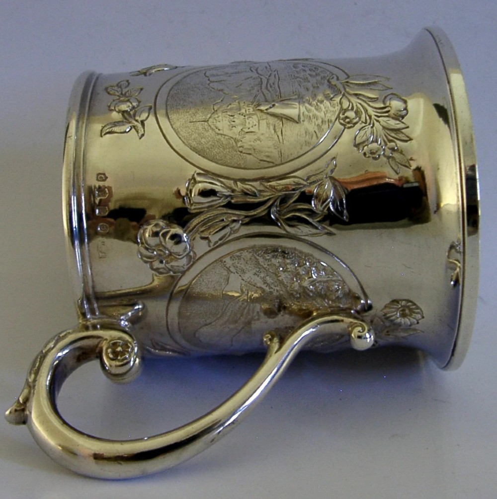 A Victorian sterling silver tankard mug featuring detailed relief floral decoration, a landscape roundel, a scroll handle, and a gilt interior. View X