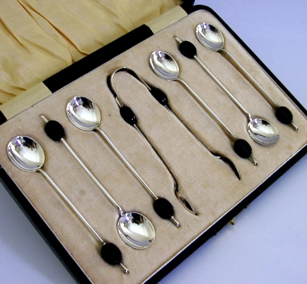 A fitted display case containing six sterling silver coffee spoons with black coffee bean finials and matching sugar tongs.