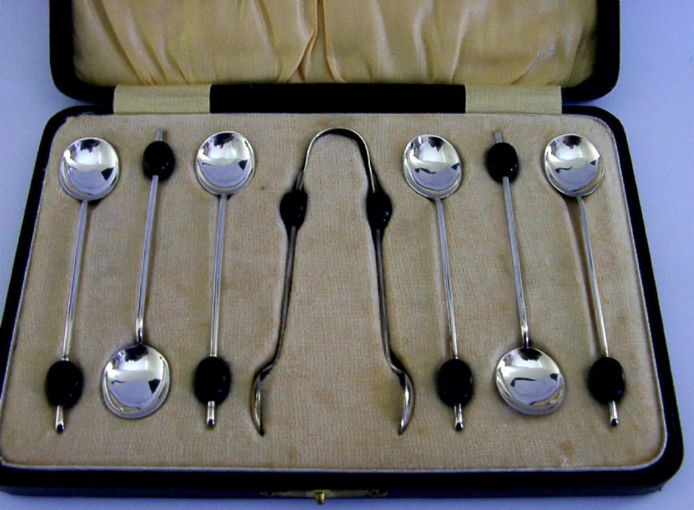 A fitted display case containing six sterling silver coffee spoons with black coffee bean finials and matching sugar tongs. View II