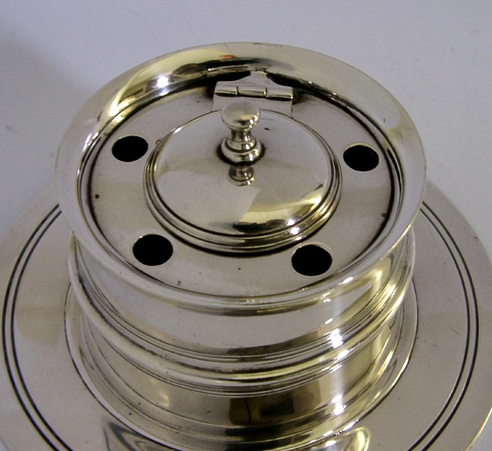 A solid sterling silver circular capstan inkwell featuring a hinged central lid and pierced quill holes set upon a wide dished base with visible hallmarks. View V