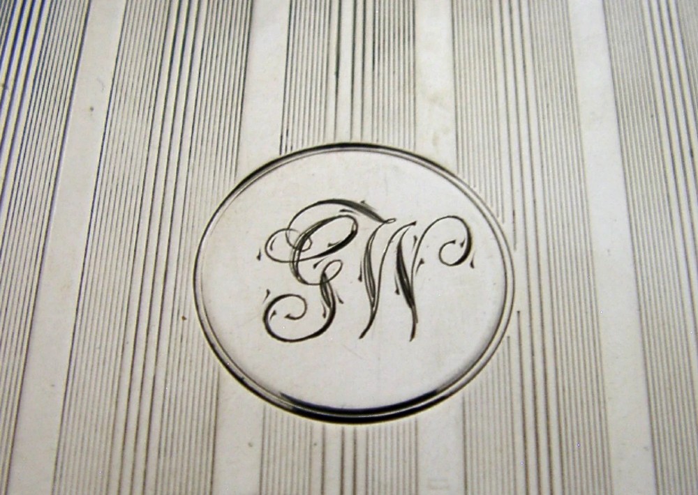 An open rectangular sterling silver card case with linear engine-turned patterns and a central circular monogrammed cartouche. View II