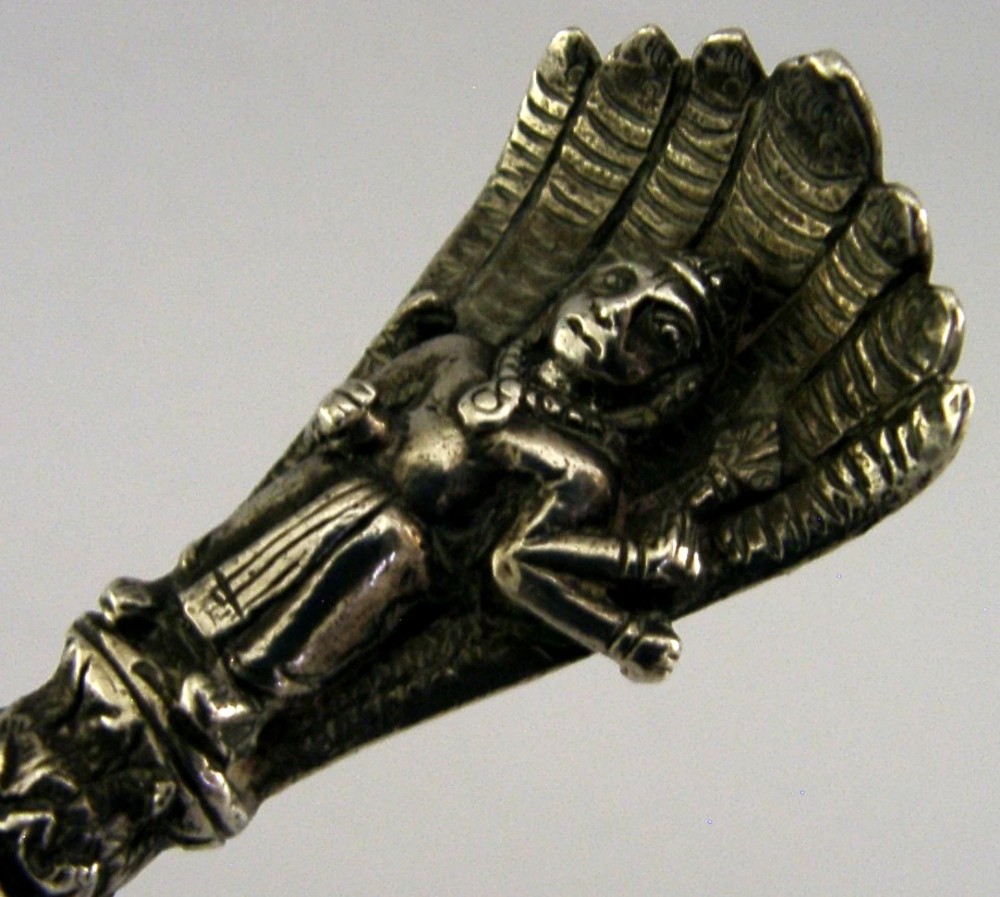 An antique silver offering spoon featuring a shell-shaped bowl, a decorated twisted stem, and a deity finial framed by a snake hood. View VI