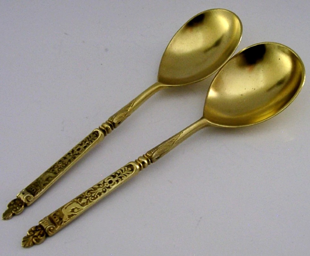 A pair of Victorian sterling silver gilt serving spoons featuring oval bowls and detailed cast handles with grotesque mask and scroll motifs.