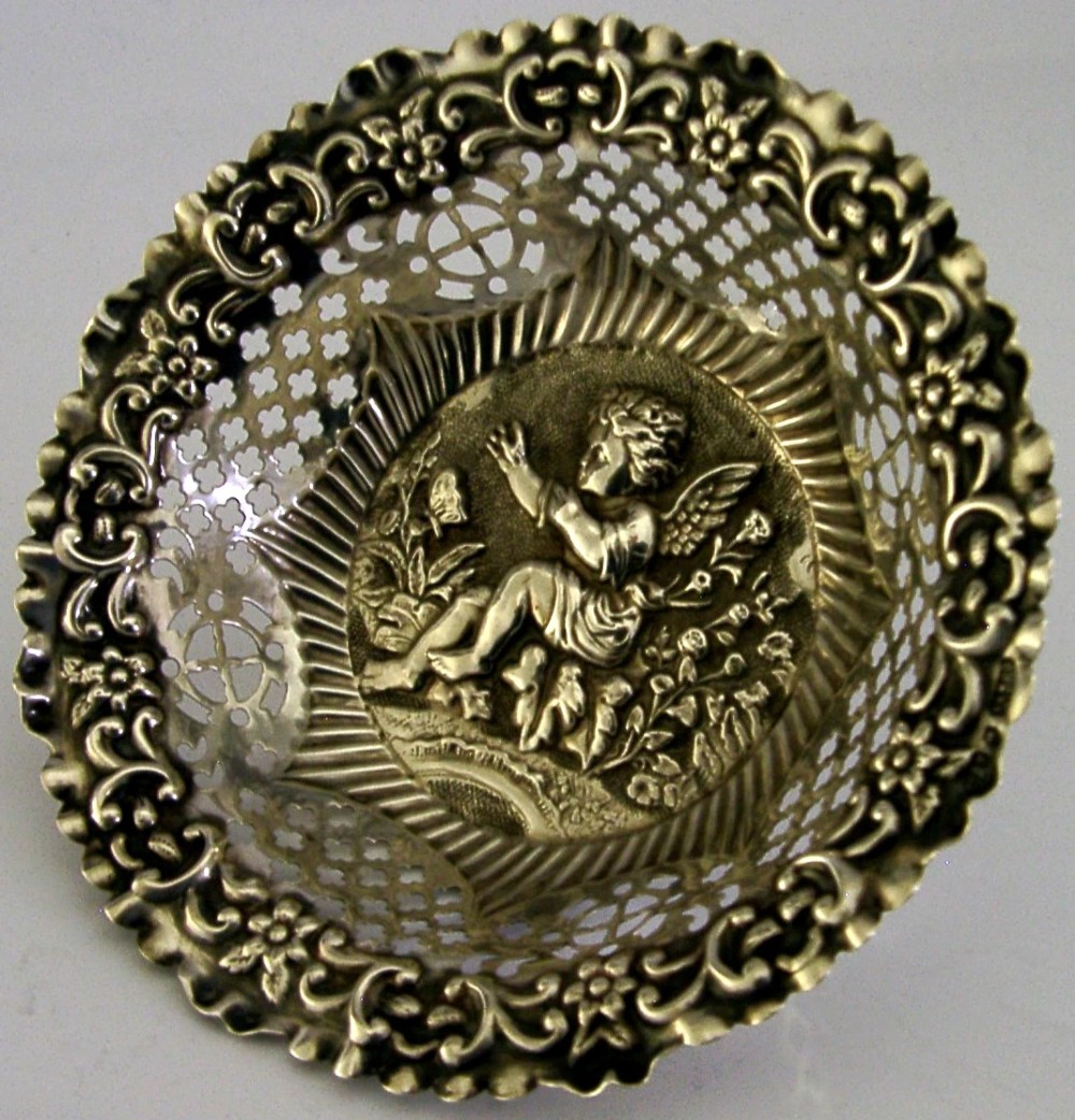 A circular sterling silver dish featuring a raised central motif of a cherub and butterfly surrounded by intricate pierced lattice work and a scalloped floral border.