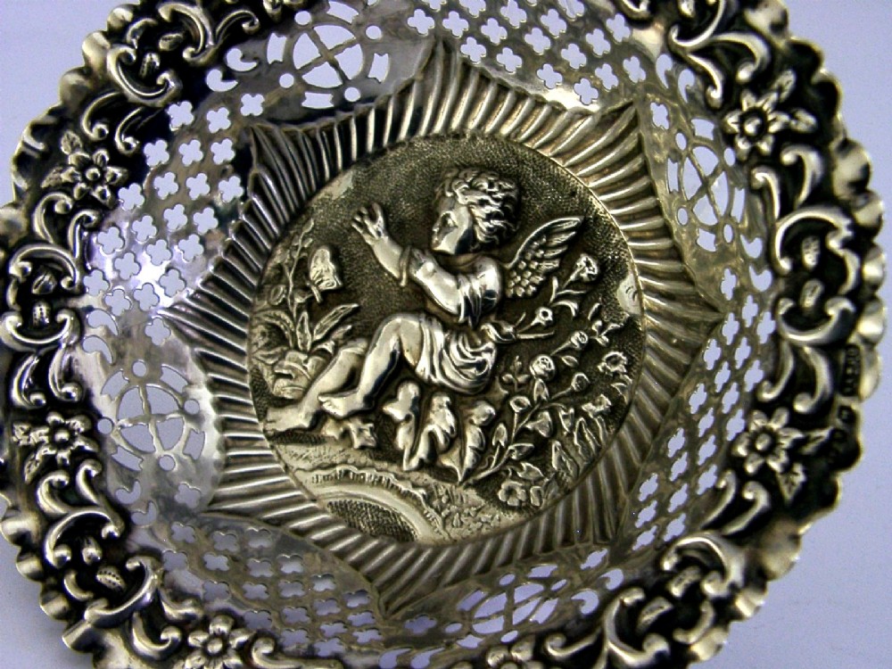 A circular sterling silver dish featuring a raised central motif of a cherub and butterfly surrounded by intricate pierced lattice work and a scalloped floral border. View III