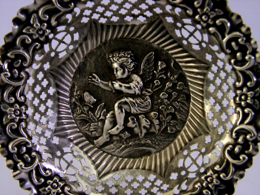 A circular sterling silver dish featuring a raised central motif of a cherub and butterfly surrounded by intricate pierced lattice work and a scalloped floral border. View IV
