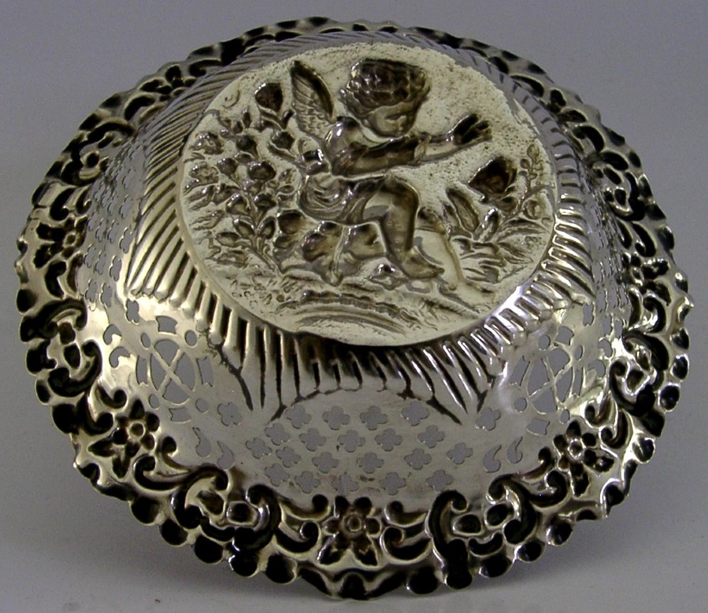 A circular sterling silver dish featuring a raised central motif of a cherub and butterfly surrounded by intricate pierced lattice work and a scalloped floral border. View V
