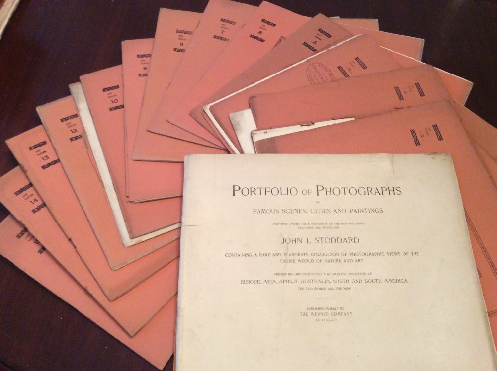 A fan-shaped arrangement of sixteen dusty pink late Victorian softcover portfolios with a main title cover reading Portfolio of Photographs by John L Stoddard.