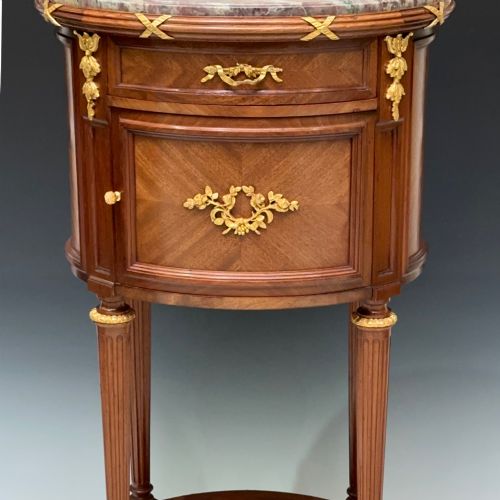 fig 1. An oval French mahogany table de nuit featuring a grey marble top, gilt-bronze mounts, single drawer, cupboard door, and fluted legs with an undertier.