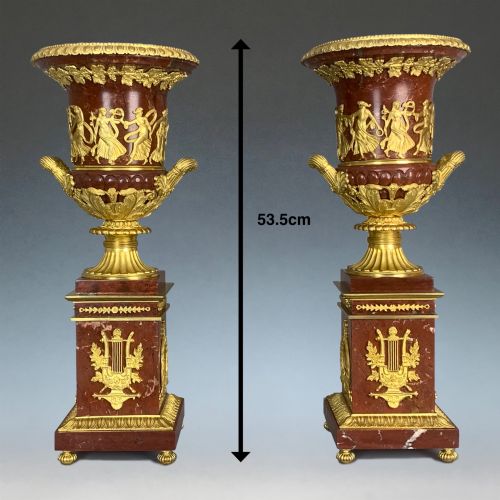 fig 1. A pair of French Empire style red marble urns featuring gilded bronze classical figure reliefs and mounted on square marble pedestals with lyre details.