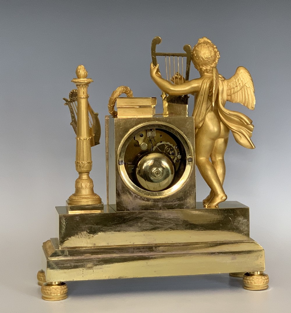A 19th-century French Empire gilt bronze mantel clock featuring a cherub figure holding a lyre alongside classical decorative elements. View VI