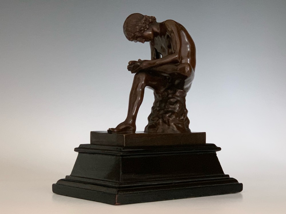 A dark patinated bronze sculpture depicting a nude youth seated on a rock, leaning forward to remove a thorn from his foot, set upon a rectangular bronze base. View II
