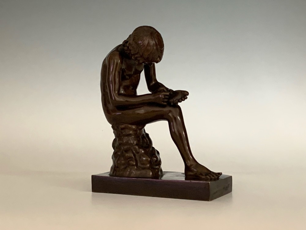 A dark patinated bronze sculpture depicting a nude youth seated on a rock, leaning forward to remove a thorn from his foot, set upon a rectangular bronze base. View III