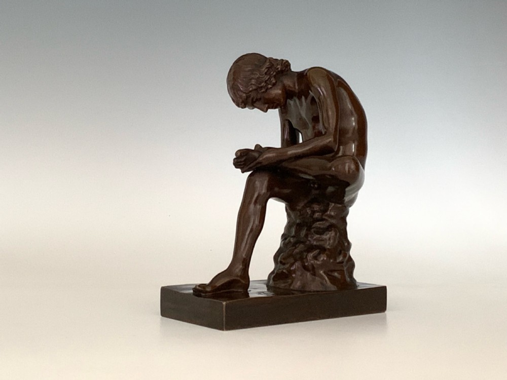 A dark patinated bronze sculpture depicting a nude youth seated on a rock, leaning forward to remove a thorn from his foot, set upon a rectangular bronze base. View IV