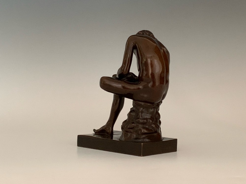 A dark patinated bronze sculpture depicting a nude youth seated on a rock, leaning forward to remove a thorn from his foot, set upon a rectangular bronze base. View V