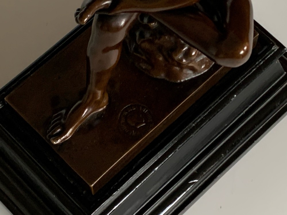 A dark patinated bronze sculpture depicting a nude youth seated on a rock, leaning forward to remove a thorn from his foot, set upon a rectangular bronze base. View VI