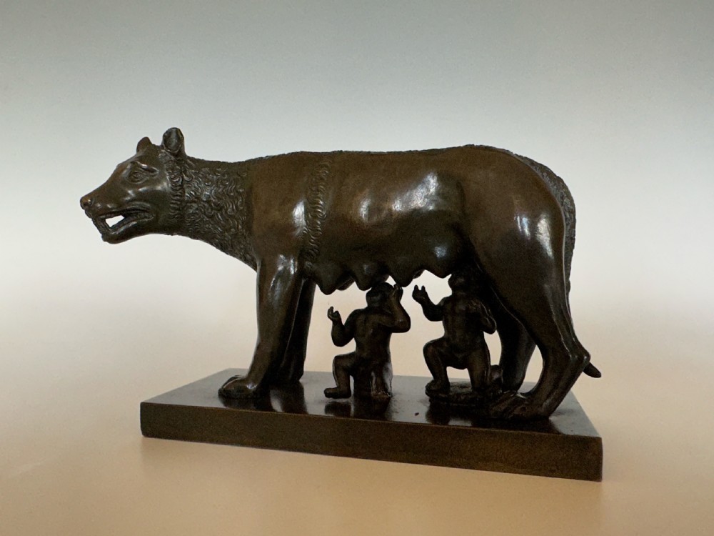 A dark bronze sculpture depicting the Capitoline Wolf nursing Romulus and Remus on a rectangular bronze base.