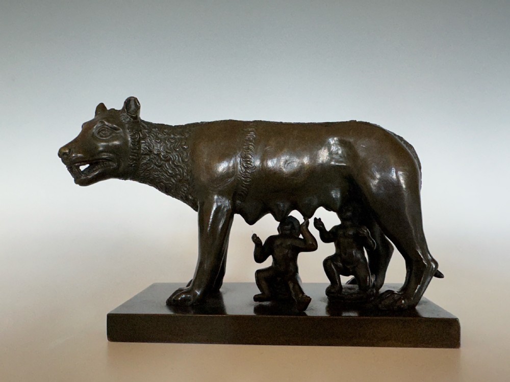 A dark bronze sculpture depicting the Capitoline Wolf nursing Romulus and Remus on a rectangular bronze base. View III