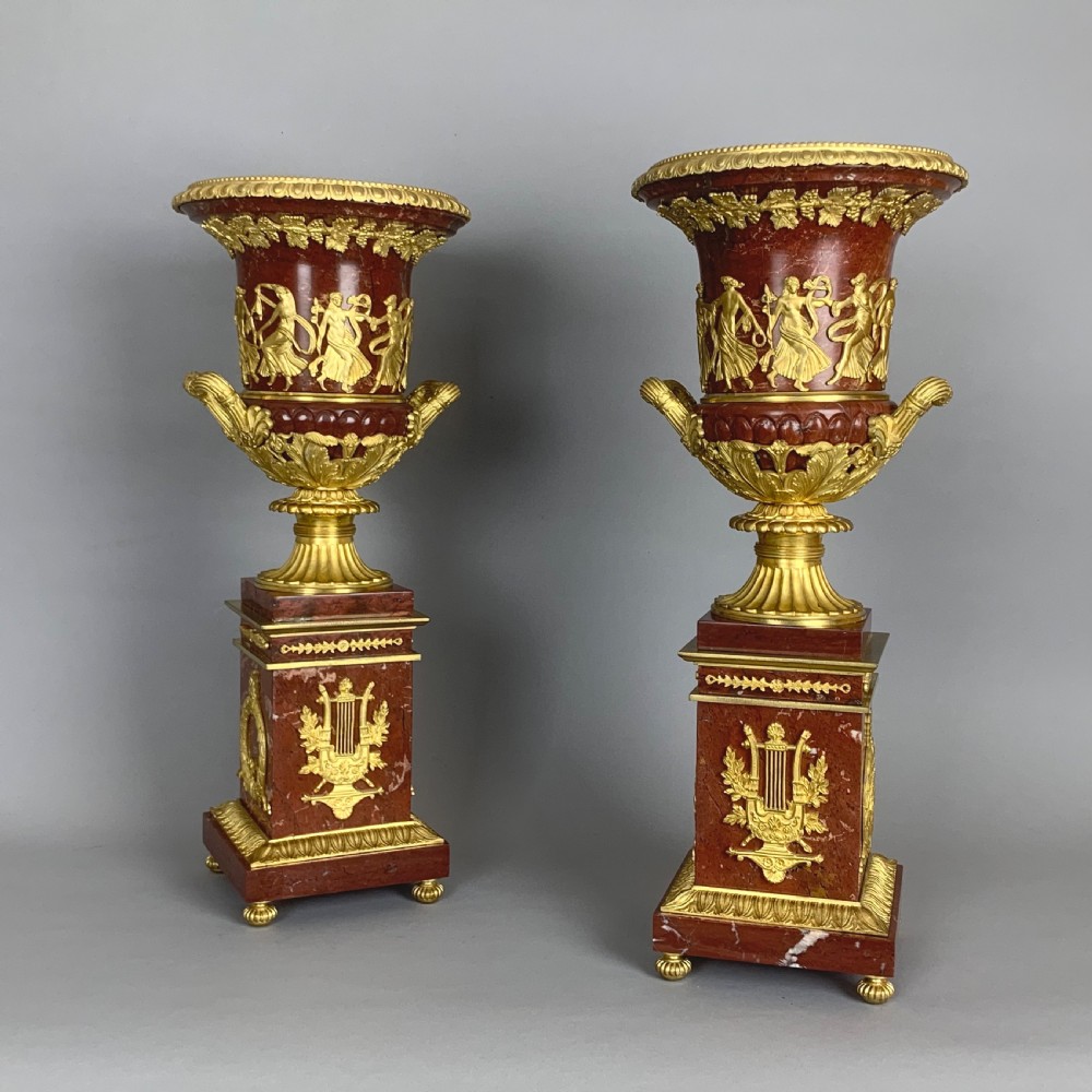 A pair of red marble urns featuring gilt bronze mounts, classical relief friezes of dancing figures, and pedestal bases decorated with lyres.