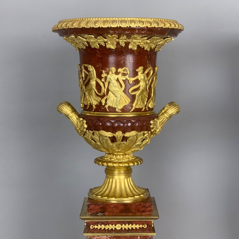 A pair of red marble urns featuring gilt bronze mounts, classical relief friezes of dancing figures, and pedestal bases decorated with lyres. View II