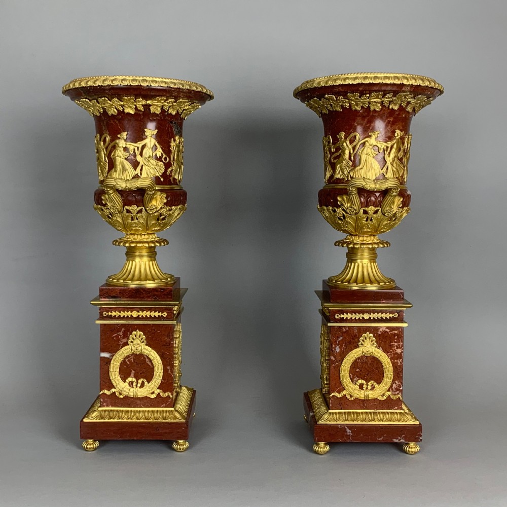 A pair of red marble urns featuring gilt bronze mounts, classical relief friezes of dancing figures, and pedestal bases decorated with lyres. View V