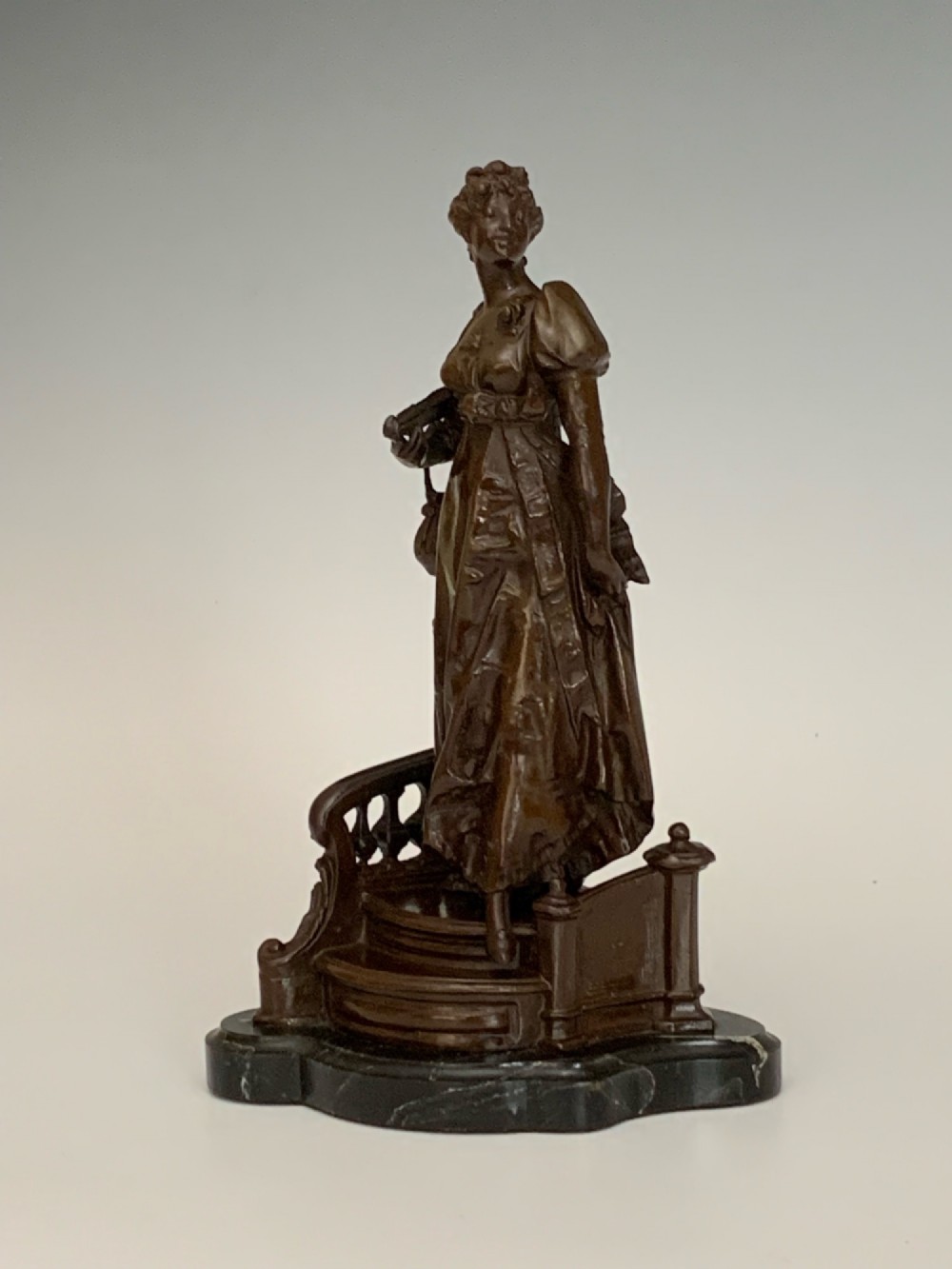 A patinated dark bronze sculpture of a woman in period dress standing on steps with a balustrade mounted on a dark marble base. View II