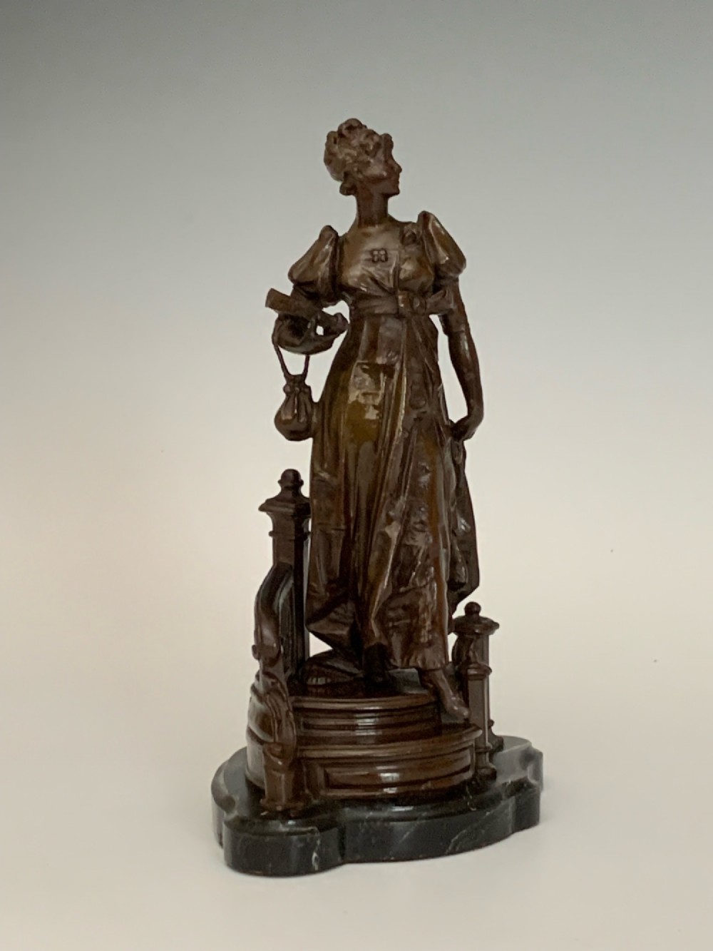 A patinated dark bronze sculpture of a woman in period dress standing on steps with a balustrade mounted on a dark marble base. View III