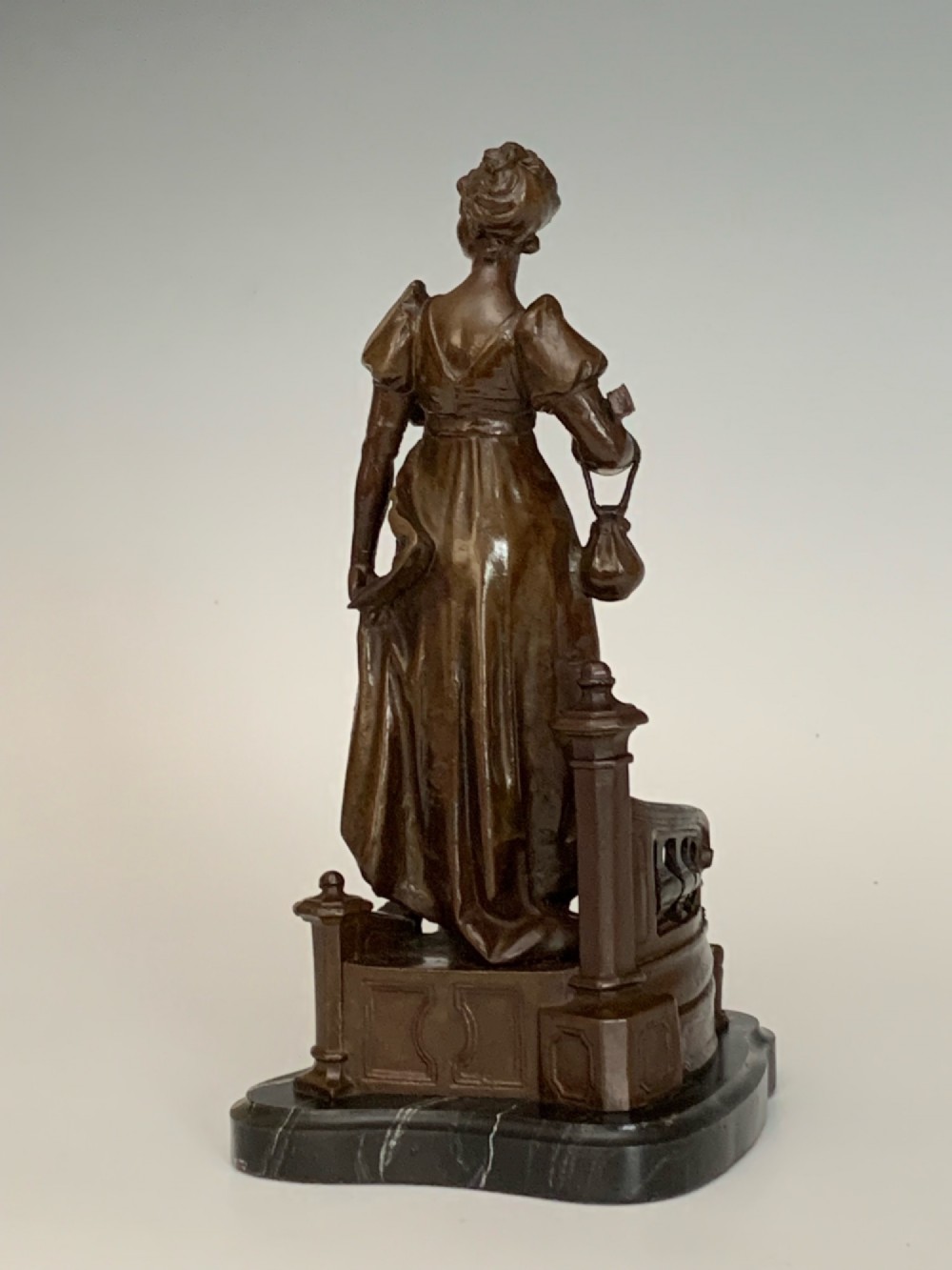 A patinated dark bronze sculpture of a woman in period dress standing on steps with a balustrade mounted on a dark marble base. View IV