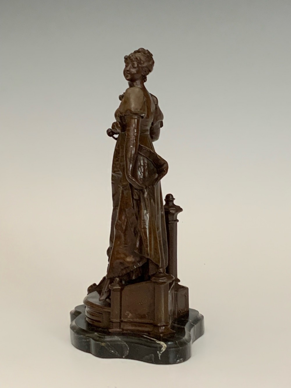 A patinated dark bronze sculpture of a woman in period dress standing on steps with a balustrade mounted on a dark marble base. View V