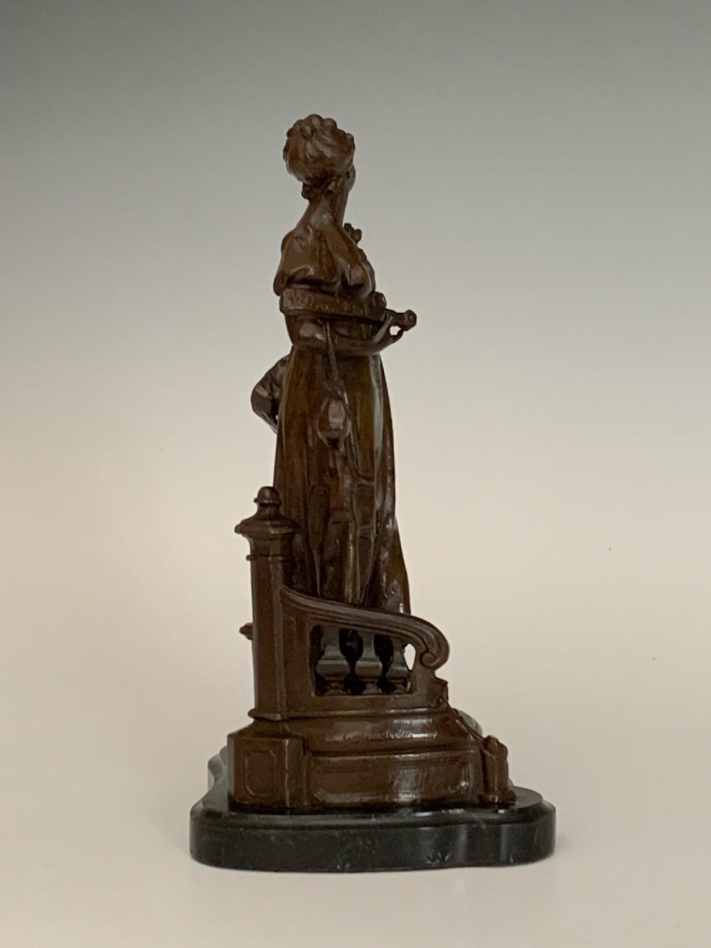 A patinated dark bronze sculpture of a woman in period dress standing on steps with a balustrade mounted on a dark marble base. View VII