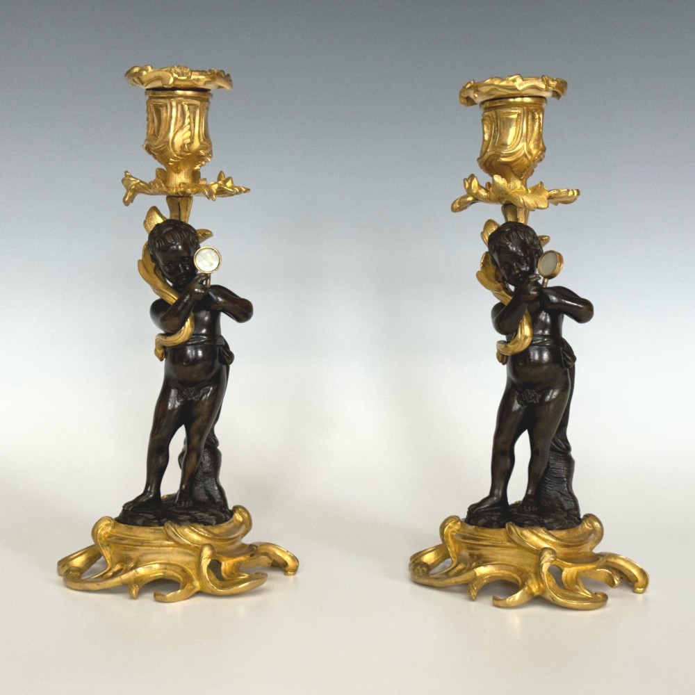 A pair of French Napoleon III candlesticks featuring dark patinated bronze cherubs standing on scrolling gilt ormolu bases supporting decorative foliate candle holders.