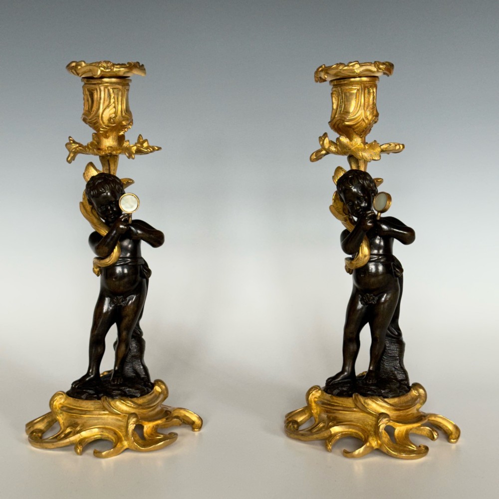 A pair of French Napoleon III candlesticks featuring dark patinated bronze cherubs standing on scrolling gilt ormolu bases supporting decorative foliate candle holders. View II