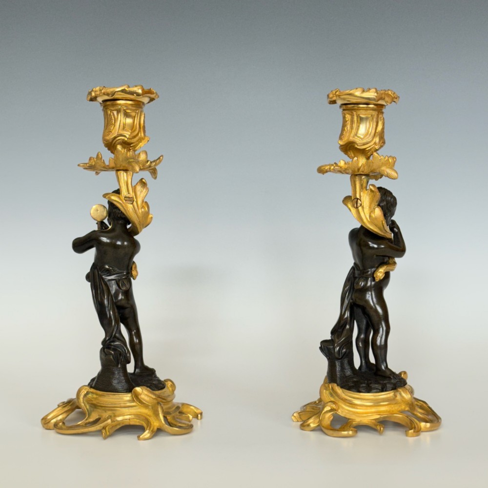 A pair of French Napoleon III candlesticks featuring dark patinated bronze cherubs standing on scrolling gilt ormolu bases supporting decorative foliate candle holders. View III