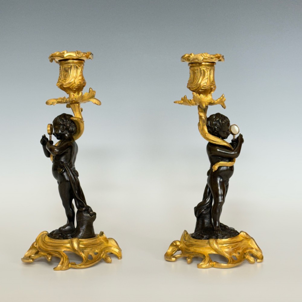 A pair of French Napoleon III candlesticks featuring dark patinated bronze cherubs standing on scrolling gilt ormolu bases supporting decorative foliate candle holders. View IV