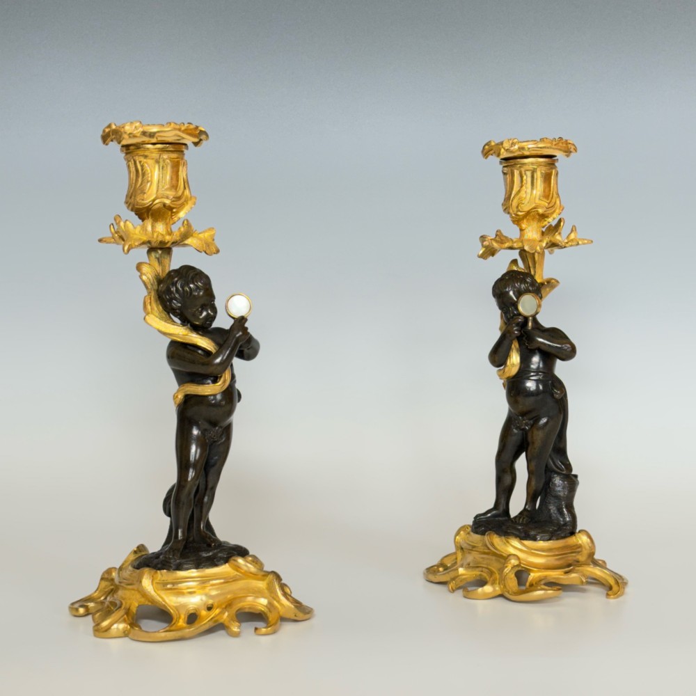 A pair of French Napoleon III candlesticks featuring dark patinated bronze cherubs standing on scrolling gilt ormolu bases supporting decorative foliate candle holders. View V