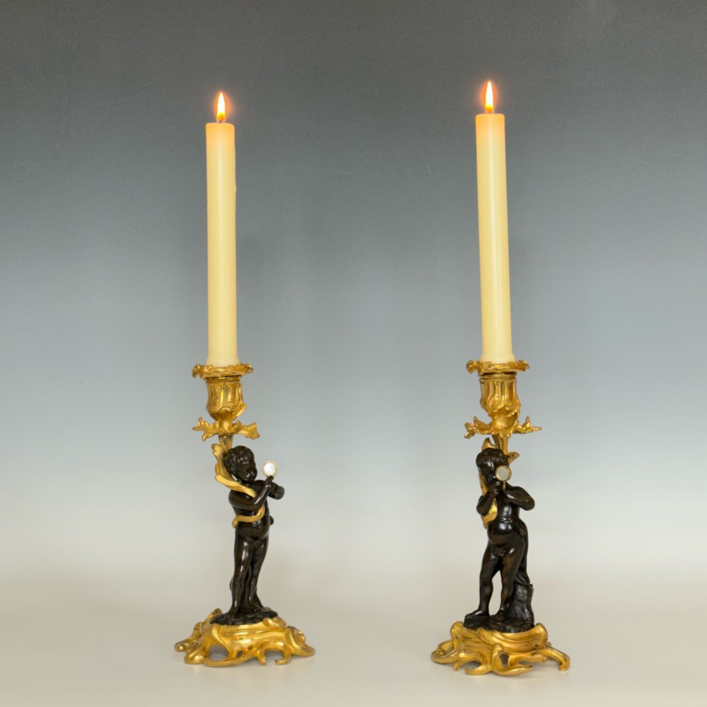 A pair of French Napoleon III candlesticks featuring dark patinated bronze cherubs standing on scrolling gilt ormolu bases supporting decorative foliate candle holders. View VI