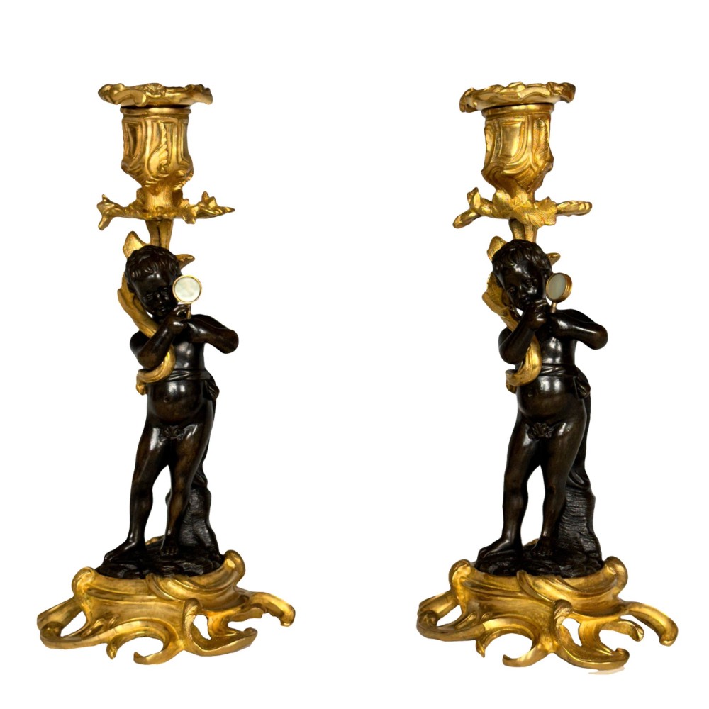 A pair of French Napoleon III candlesticks featuring dark patinated bronze cherubs standing on scrolling gilt ormolu bases supporting decorative foliate candle holders. View VIII