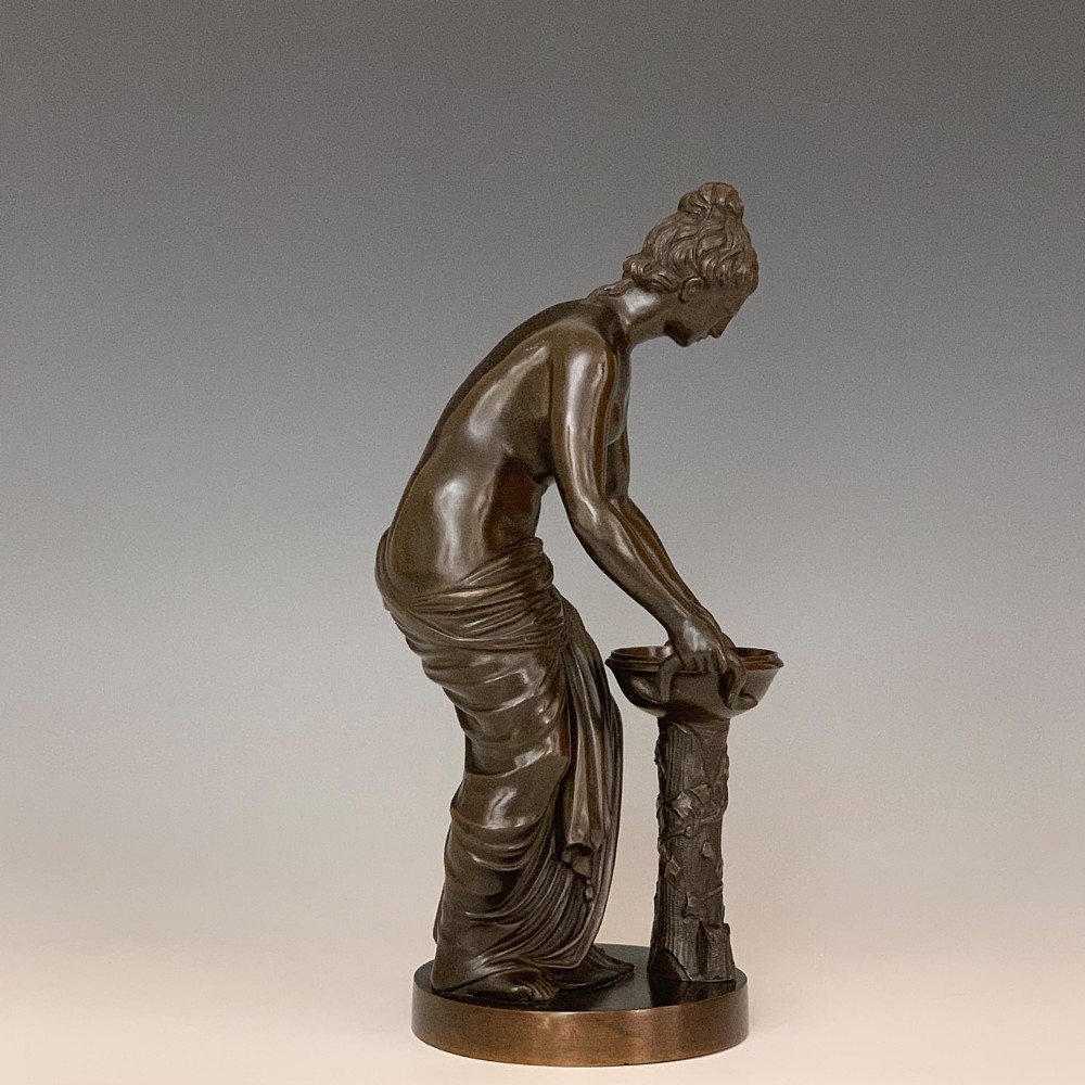 A dark brown patinated bronze figure depicting a draped female classical figure leaning over a small pedestal basin, mounted on a round base.