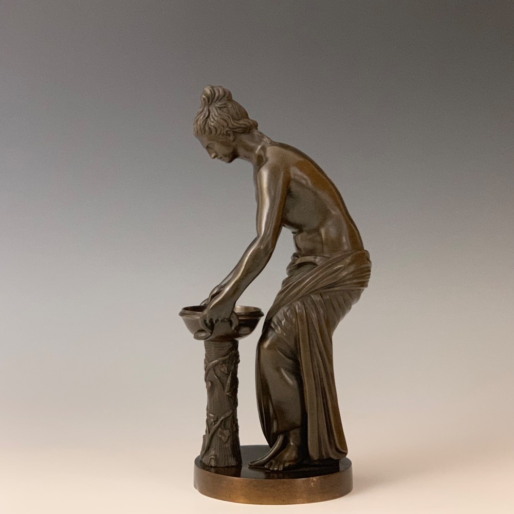 A dark brown patinated bronze figure depicting a draped female classical figure leaning over a small pedestal basin, mounted on a round base. View II