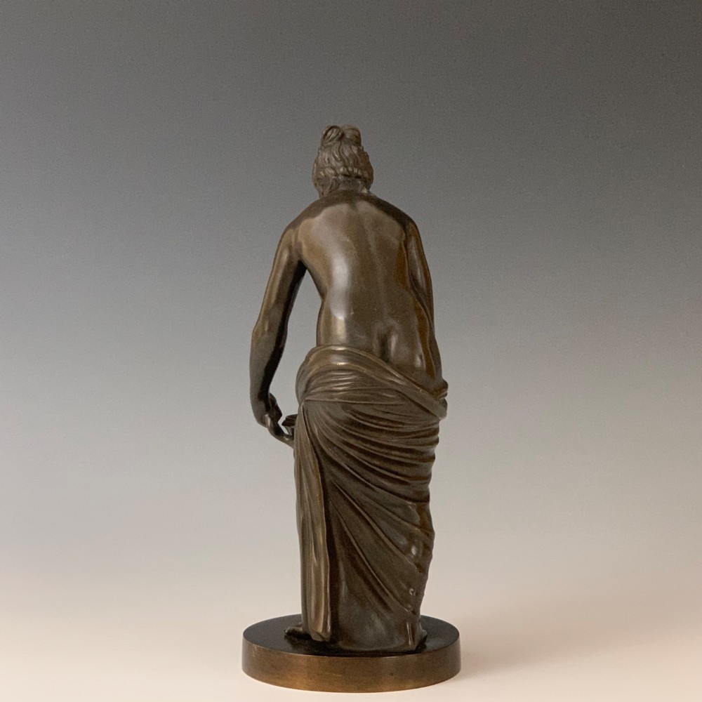 A dark brown patinated bronze figure depicting a draped female classical figure leaning over a small pedestal basin, mounted on a round base. View III