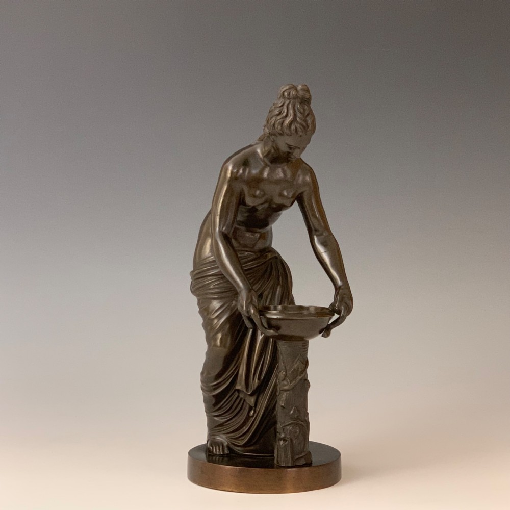 A dark brown patinated bronze figure depicting a draped female classical figure leaning over a small pedestal basin, mounted on a round base. View IV