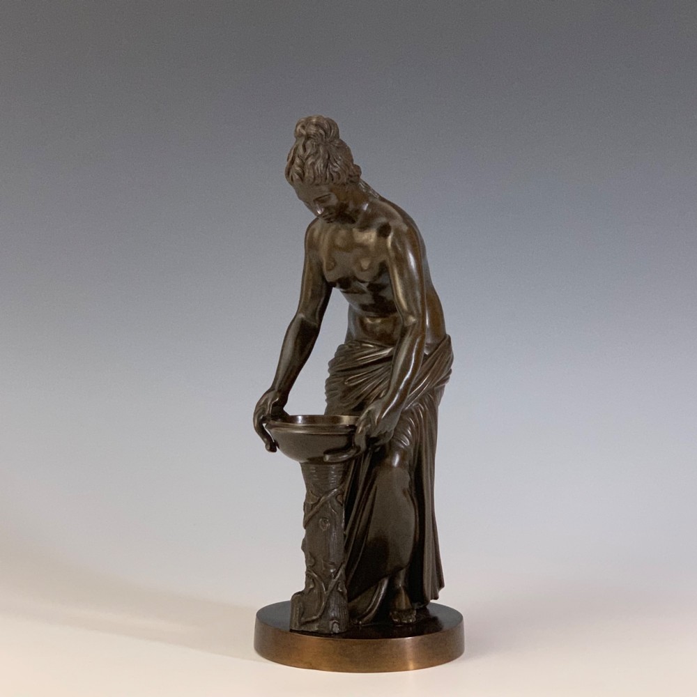A dark brown patinated bronze figure depicting a draped female classical figure leaning over a small pedestal basin, mounted on a round base. View V
