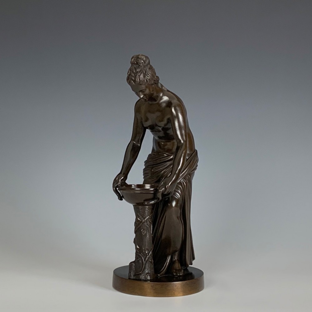 A dark brown patinated bronze figure depicting a draped female classical figure leaning over a small pedestal basin, mounted on a round base. View VI
