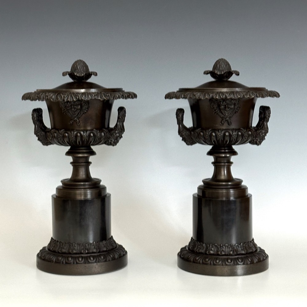A pair of patinated dark bronze covered urns featuring classical mask motifs, acanthus leaf moulding, twin handles, and pinecone finials on round stepped bases.