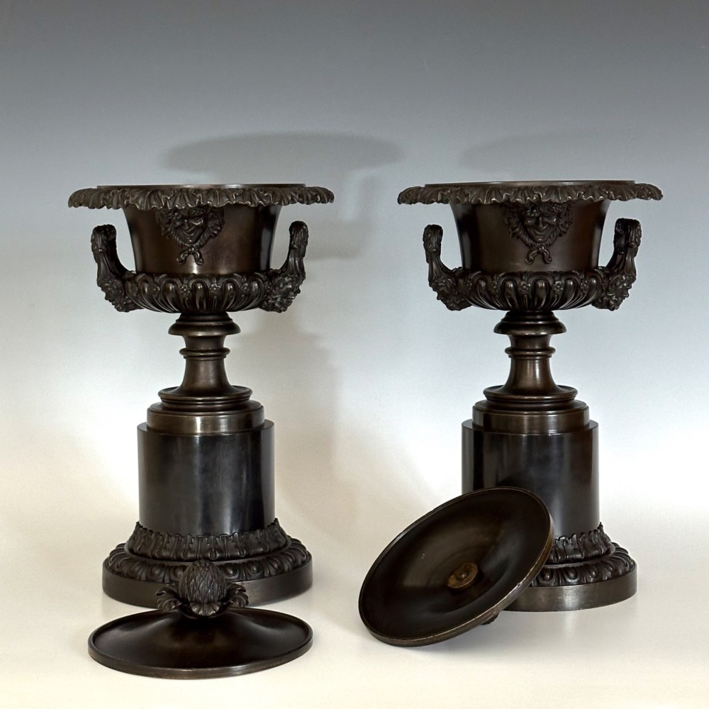 A pair of patinated dark bronze covered urns featuring classical mask motifs, acanthus leaf moulding, twin handles, and pinecone finials on round stepped bases. View II