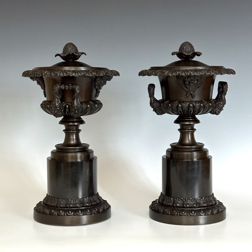 A pair of patinated dark bronze covered urns featuring classical mask motifs, acanthus leaf moulding, twin handles, and pinecone finials on round stepped bases. View III