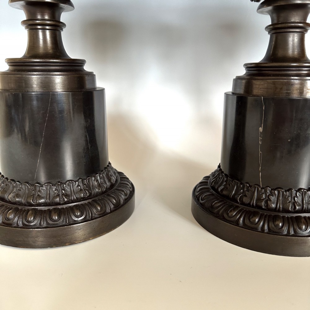 A pair of patinated dark bronze covered urns featuring classical mask motifs, acanthus leaf moulding, twin handles, and pinecone finials on round stepped bases. View IV